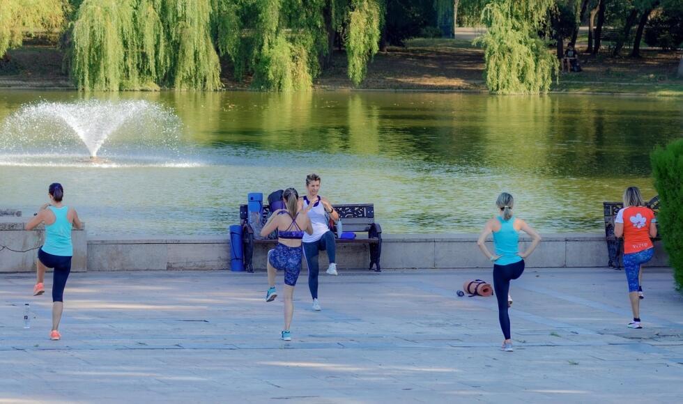 Trainer που γυμνάζει γκρουπ γυναικών σε πάρκο, μπροστά από συντριβάνι.