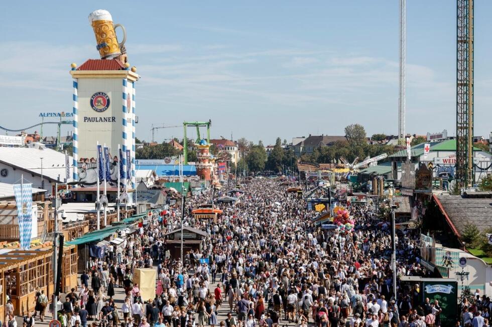 Πανοραμική άποψη του Oktoberfest στο Μόναχο με πλήθη κόσμου να περπατούν ανάμεσα σε παραδοσιακά περίπτερα, με τεράστιο διαφημιστικό πύργο μπύρας Paulaner στο αριστερό μέρος και λούνα παρκ στο βάθος